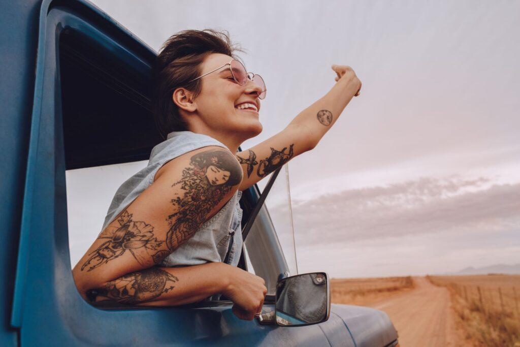 Frau mit Tattoos, die aus dem Fenster eines blauen Autos schaut, lächelnd und in Richtung Horizont zeigend, während sie eine Reise genießt.