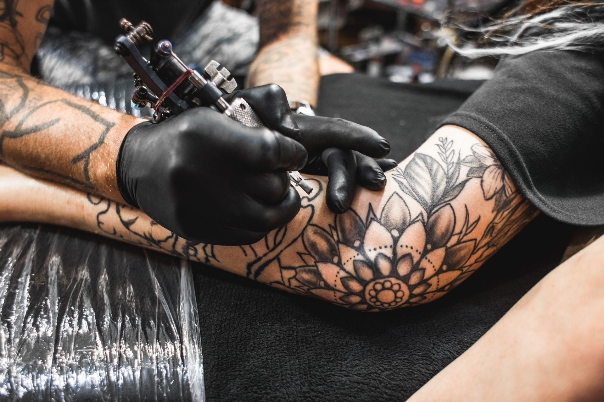 Nahaufnahme eines Tätowierers, der mit schwarzen Handschuhen ein Blumen-Tattoo auf den Arm eines Kunden sticht.